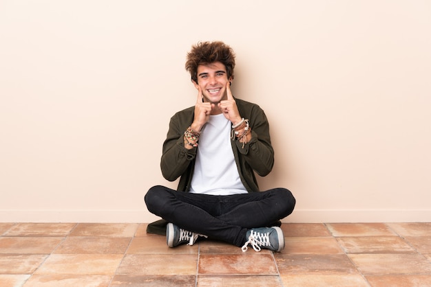 Hombre caucásico joven sentado en el suelo sonriendo con una expresión feliz y agradable