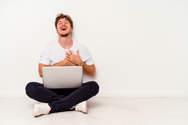 Hombre caucásico joven sentado en el suelo sosteniendo portátil aislado sobre fondo blanco riendo manteniendo las manos en el corazón, concepto de felicidad.