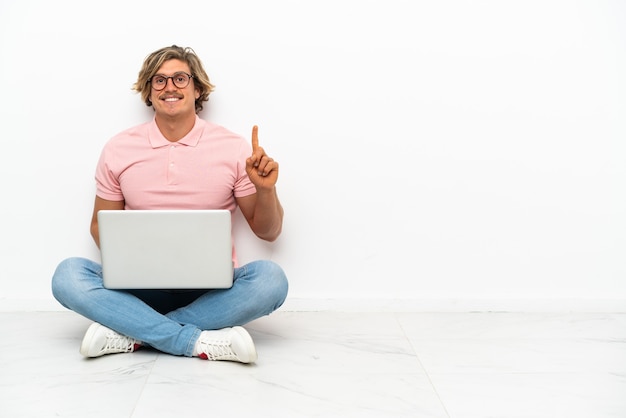 Hombre caucásico joven sentado en el suelo con su computadora portátil aislada en la pared blanca apuntando hacia una gran idea