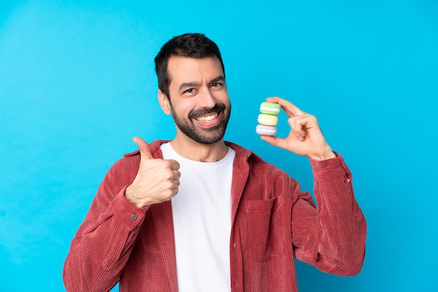 Hombre caucásico joven sobre azul aislado con coloridos macarons franceses con los pulgares para arriba