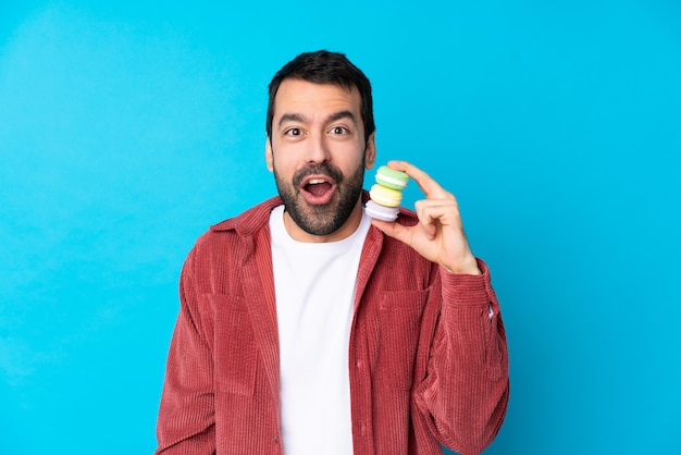 Hombre caucásico joven sobre la pared azul aislada con coloridos macarons franceses con expresión de sorpresa
