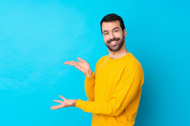 Hombre caucásico joven sobre la pared azul aislada que extiende las manos a un lado para invitar a venir