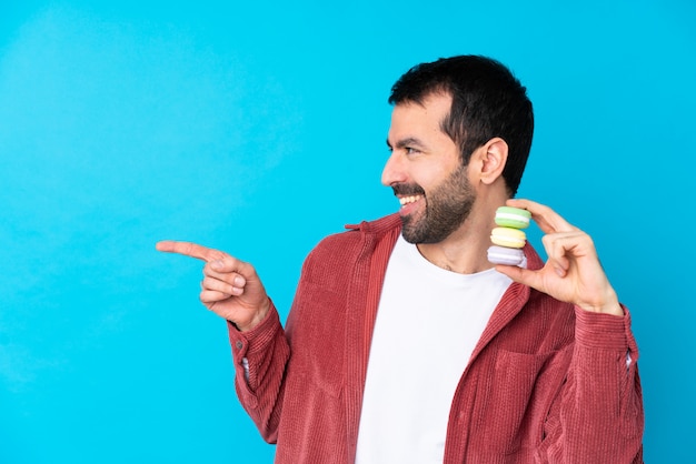 Hombre caucásico joven sobre la pared azul aislada que sostiene los macarons franceses coloridos y que señala el lado