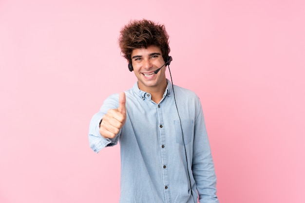 Hombre caucásico joven sobre la pared rosada aislada que trabaja con el auricular con el pulgar para arriba