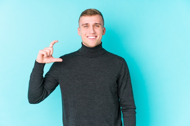 Hombre caucásico joven sobre una superficie azul sosteniendo algo pequeño con los dedos índices, sonriente y confiado.