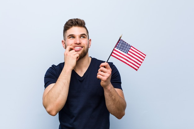 Hombre caucásico joven sosteniendo una bandera de Estados Unidos relajado pensando en algo