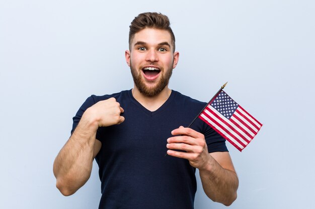 Hombre caucásico joven sosteniendo una bandera de Estados Unidos sorprendido señalando a sí mismo, sonriendo ampliamente.