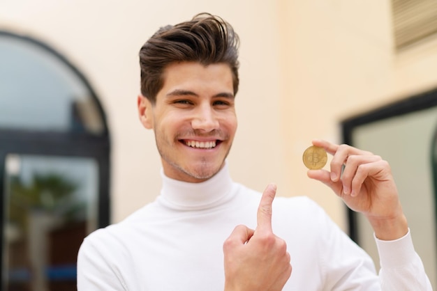 Hombre caucásico joven sosteniendo un Bitcoin al aire libre y apuntándolo