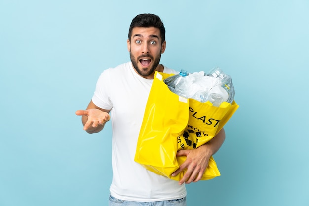 Hombre caucásico joven sosteniendo una bolsa llena de botellas de plástico para reciclar aislado sobre fondo azul con expresión facial sorprendida