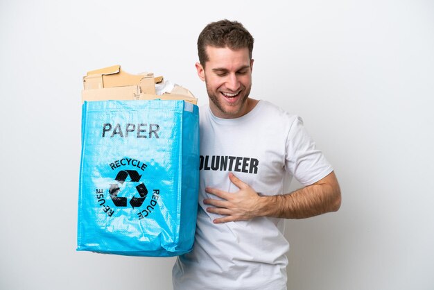 Hombre caucásico joven sosteniendo una bolsa de reciclaje llena de papel para reciclar aislado sobre fondo blanco sonriendo mucho