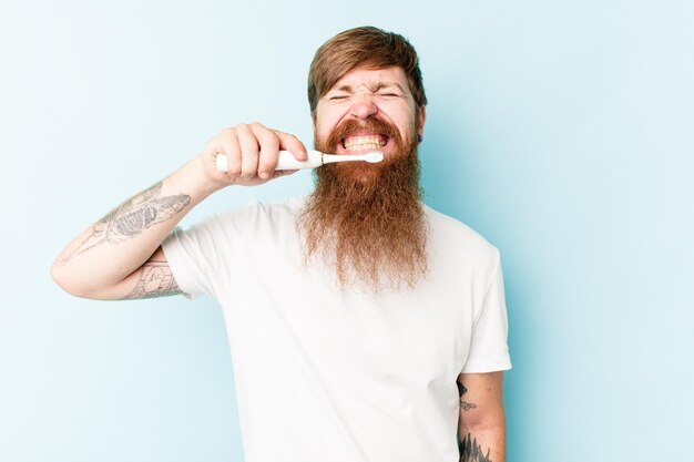 Hombre caucásico joven sosteniendo un cepillo de dientes eléctrico aislado sobre fondo azul.