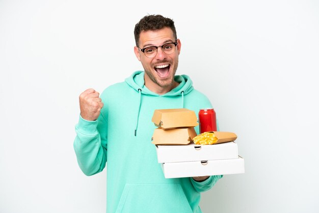 Hombre caucásico joven sosteniendo comida rápida aislado sobre fondo blanco celebrando una victoria en posición de ganador