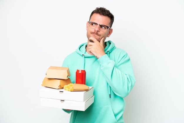 Hombre caucásico joven sosteniendo comida rápida aislado sobre fondo blanco que tiene dudas y pensamiento