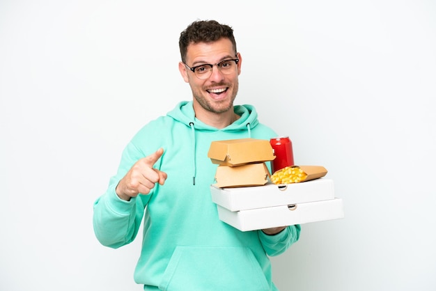 Hombre caucásico joven sosteniendo comida rápida aislado sobre fondo blanco sorprendido y apuntando al frente