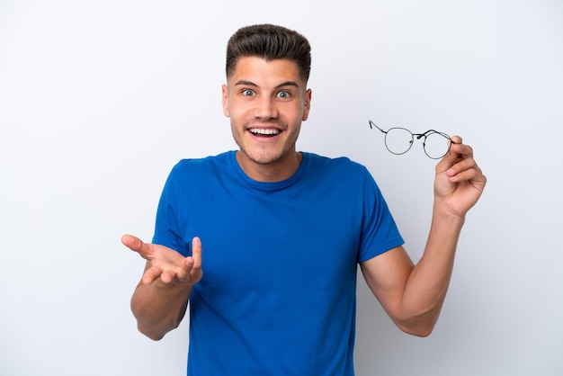 Hombre caucásico joven sosteniendo gafas aislado sobre fondo blanco con expresión facial sorprendida