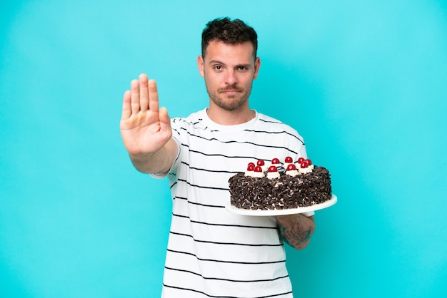 Hombre caucásico joven sosteniendo pastel de cumpleaños aislado sobre fondo azul haciendo gesto de parada