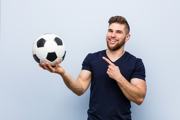 Hombre caucásico joven sosteniendo una pelota de fútbol sonriendo alegremente apuntando con el dedo lejos.