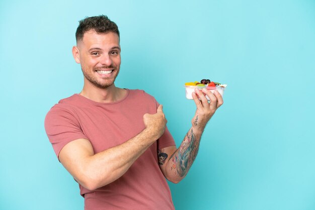 Hombre caucásico joven sosteniendo un plato de fruta aislado sobre fondo azul apuntando hacia atrás