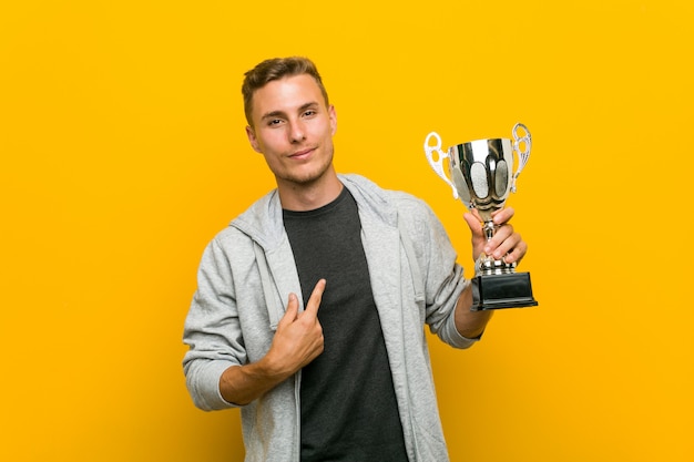 Hombre caucásico joven sosteniendo un trofeo apuntando con el dedo hacia usted como si invitara a acercarse.