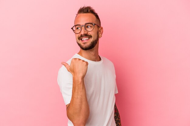 Hombre caucásico joven con tatuajes aislados en puntos de fondo rosa con el dedo pulgar lejos, riendo y despreocupado.