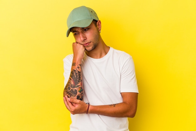 Hombre caucásico joven con tatuajes aislados sobre fondo amarillo que se siente triste y pensativo, mirando el espacio de la copia.