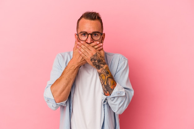 Hombre caucásico joven con tatuajes aislados sobre fondo rosa cubriendo la boca con las manos mirando preocupado.