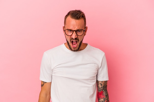 Hombre caucásico joven con tatuajes aislados sobre fondo rosa gritando muy enojado, concepto de rabia, frustrado.