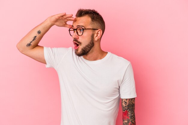 Hombre caucásico joven con tatuajes aislados sobre fondo rosa mirando lejos manteniendo la mano en la frente.