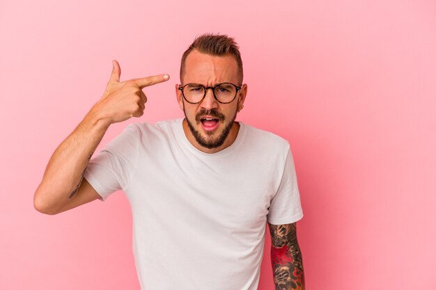 Hombre caucásico joven con tatuajes aislados sobre fondo rosa mostrando un gesto de decepción con el dedo índice.