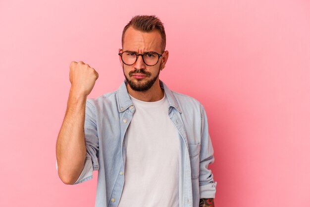 Hombre caucásico joven con tatuajes aislados sobre fondo rosa mostrando el puño a la cámara, expresión facial agresiva.