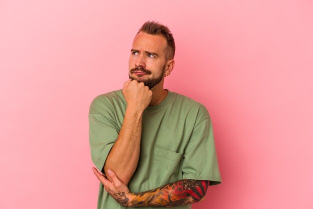 Hombre caucásico joven con tatuajes aislados sobre fondo rosa relajado pensando en algo mirando un espacio de copia.