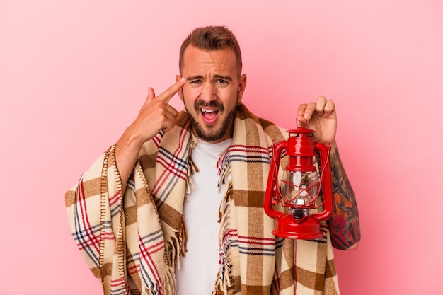 Hombre caucásico joven con tatuajes sosteniendo linterna vintage aislado sobre fondo rosa mostrando un gesto de decepción con el dedo índice.