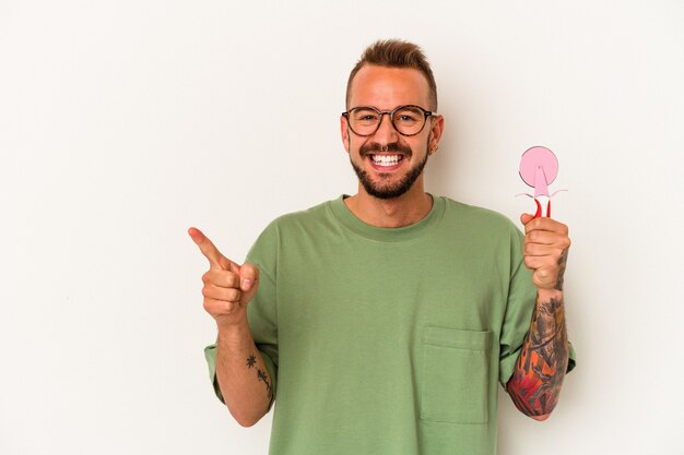 Hombre caucásico joven con tatuajes sosteniendo piruleta aislado sobre fondo blanco sonriendo y apuntando a un lado, mostrando algo en el espacio en blanco.