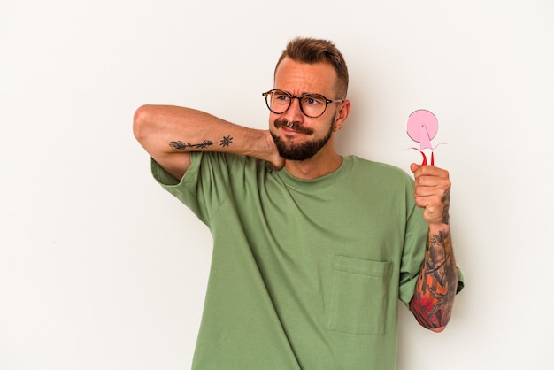 Hombre caucásico joven con tatuajes sosteniendo piruleta aislado sobre fondo blanco tocando la parte posterior de la cabeza, pensando y haciendo una elección.