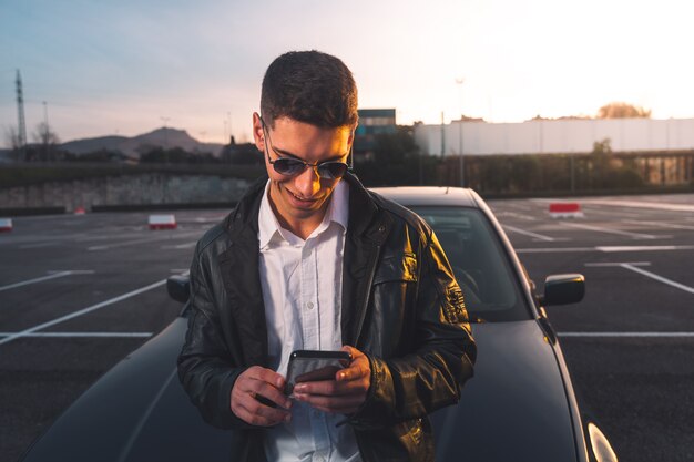 Hombre caucásico joven con un teléfono inteligente con un coche deportivo detrás