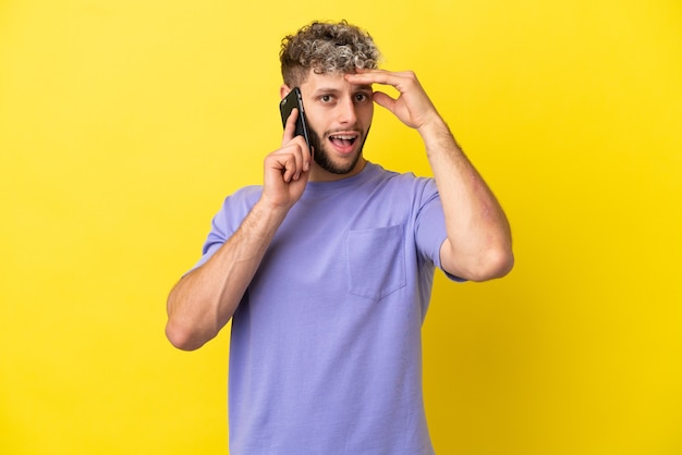 Hombre caucásico joven con teléfono móvil aislado sobre fondo amarillo haciendo gesto de sorpresa mientras mira hacia el lado