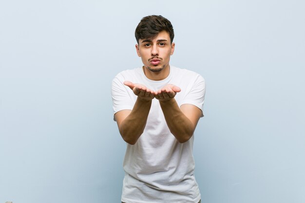 Hombre caucásico joven vistiendo una camiseta blanca doblando los labios y sosteniendo las palmas para enviar beso de aire.