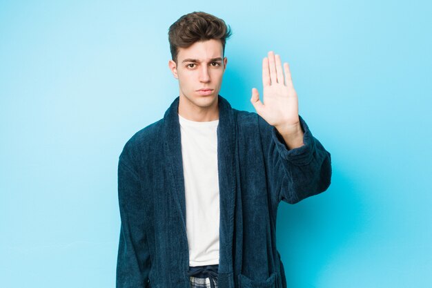 Hombre caucásico joven vistiendo pijamas de pie con la mano extendida que muestra la señal de stop, impidiéndole.