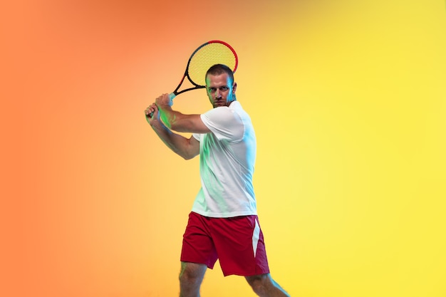 Un hombre caucásico jugando al tenis aislado sobre fondo de estudio en luz de neón. Colocar al joven jugador profesional masculino en movimiento o acción durante el juego deportivo. Concepto de movimiento, deporte, estilo de vida saludable.