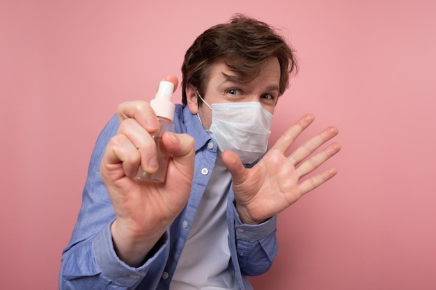 Hombre caucásico con máscara médica rociando spray desinfectante sobre fondo rosa