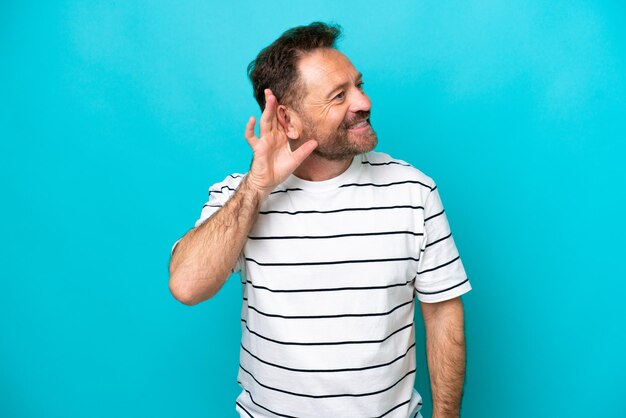 Hombre caucásico de mediana edad aislado de fondo azul escuchando algo poniendo la mano en la oreja