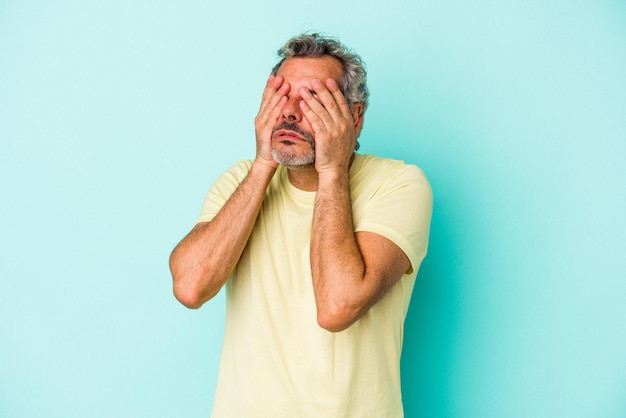 Hombre caucásico de mediana edad aislado de fondo azul con miedo cubriendo los ojos con las manos