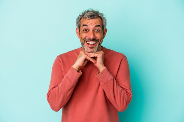 Hombre caucásico de mediana edad aislado de fondo azul rezando por suerte asombrado y abriendo la boca mirando al frente