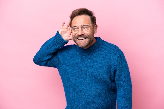 Hombre caucásico de mediana edad aislado de fondo rosa escuchando algo poniendo la mano en la oreja