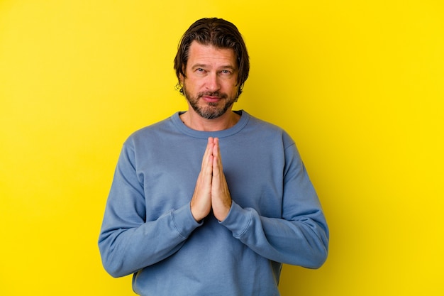 Hombre caucásico de mediana edad aislado en la pared amarilla rezando, mostrando devoción, persona religiosa en busca de inspiración divina.