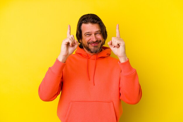 Hombre caucásico de mediana edad aislado sobre fondo amarillo indica con ambos dedos índice hacia arriba mostrando un espacio en blanco.