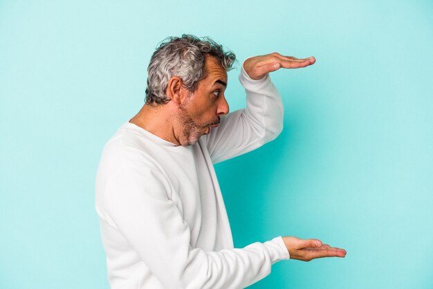 Hombre caucásico de mediana edad aislado sobre fondo azul conmocionado y sorprendido sosteniendo un espacio de copia entre las manos.