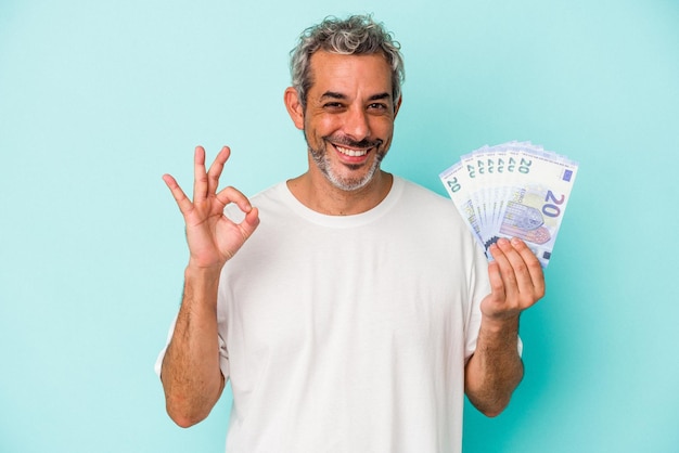 Hombre caucásico de mediana edad con billetes aislados de fondo azul alegre y confiado mostrando un gesto correcto.