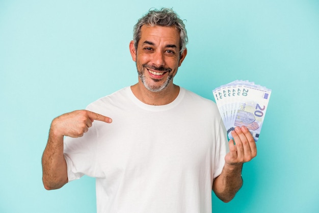 Hombre caucásico de mediana edad con billetes aislados en una persona de fondo azul que señala con la mano un espacio de copia de camisa orgulloso y confiado