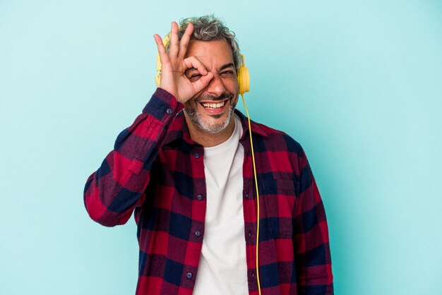 Hombre caucásico de mediana edad escuchando música aislado de fondo azul emocionado manteniendo el gesto correcto en el ojo